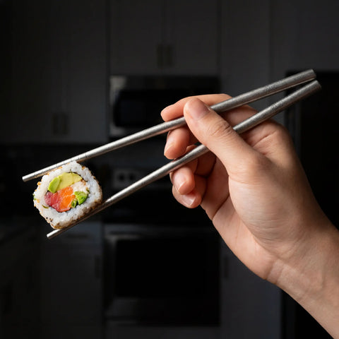 Hand holding a sushi roll with PureTi titanium chopsticks in a kitchen