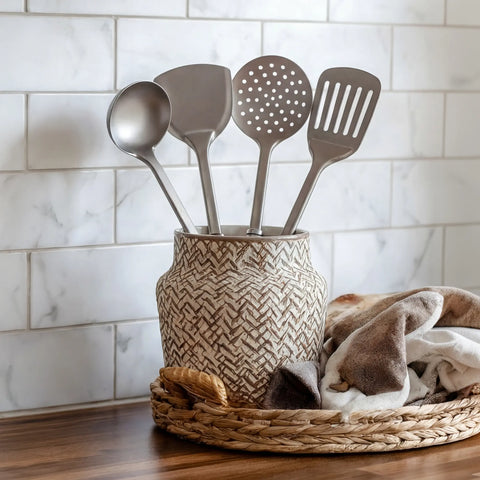Four PureTi titanium utensils in a patterned ceramic crock on a wooden bench against white tiles
