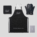 PureTi Signature Kitchen Set: black apron, silicone oven mitts, waffle weave towel and gift box on a light gray background