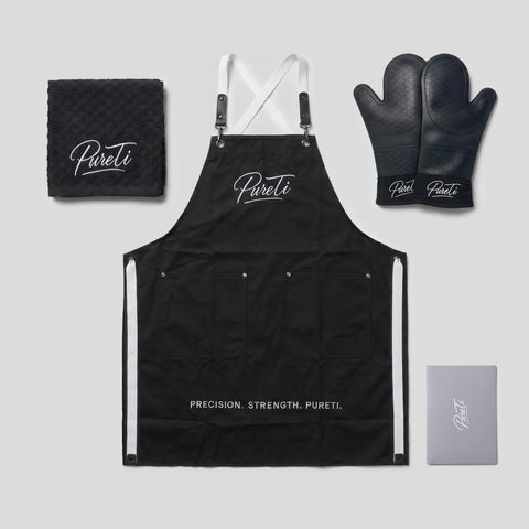 PureTi Signature Kitchen Set: black apron, silicone oven mitts, waffle weave towel and gift box on a light gray background