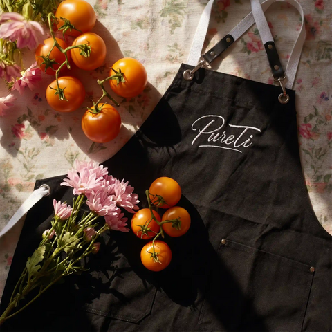 Black apron with 'PureTi' branding, tomatoes, and flowers on a floral tablecloth.