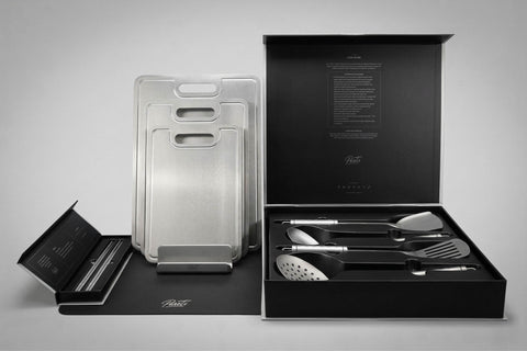 Luxury PureTi kitchenware set with titanium cutting boards, utensils in presentation boxes, a silicone mat with a silver PureTi logo, and four titanium chopsticks in a black box.