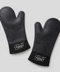 Pair of black PureTi silicone oven mitts with embroidered logo on a light gray background