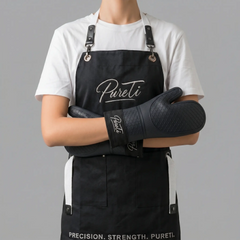 Person wearing a black apron with 'Pureti' branding and holding oven mitts against a gray background.